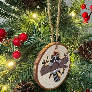 Op de afbeelding: Een houten kerstversiering met een handgeschilderde wasbeer die op een tak zit met een rode kardinaal erboven. De versiering is rond en heeft een rustieke houtnerf.