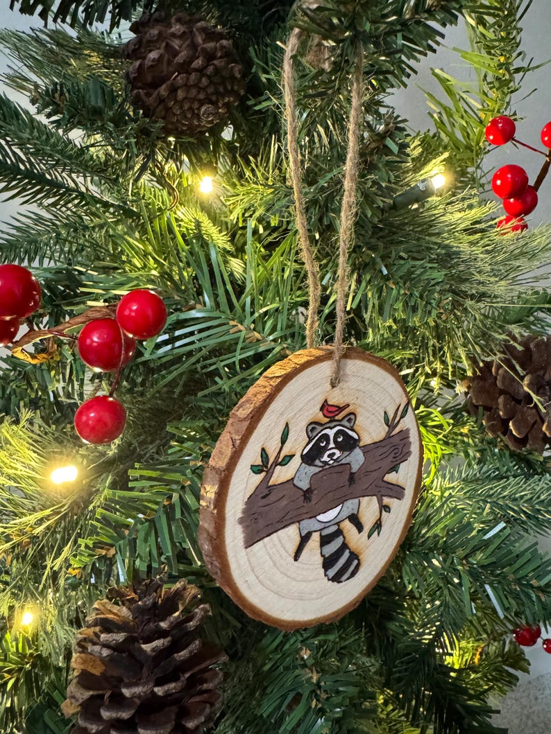 Op de afbeelding: Een houten kerstversiering met een handgeschilderde wasbeer die op een tak zit met een rode kardinaal erboven. De versiering is rond en heeft een rustieke houtnerf.