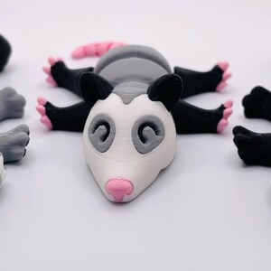 Op de afbeelding: Drie kleine, handgeschilderde dierfiguren: een wasbeer, een opossum en een stinkdier. De wasbeer is grijs met zwarte markeringen, de opossum is grijs en wit, en het stinkdier is zwart en wit.
