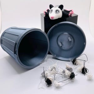 Op de afbeelding: Een blauwe plastic prullenbak met een deksel, open en leeg. Een klein zwart-wit opossum beeldje zit op het deksel. Er liggen verschillende kleine balletjes van witte en zwarte draad rond de prullenbak.