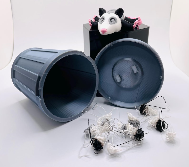 Op de afbeelding: Een blauwe plastic prullenbak met een deksel, open en leeg. Een klein zwart-wit opossum beeldje zit op het deksel. Er liggen verschillende kleine balletjes van witte en zwarte draad rond de prullenbak.