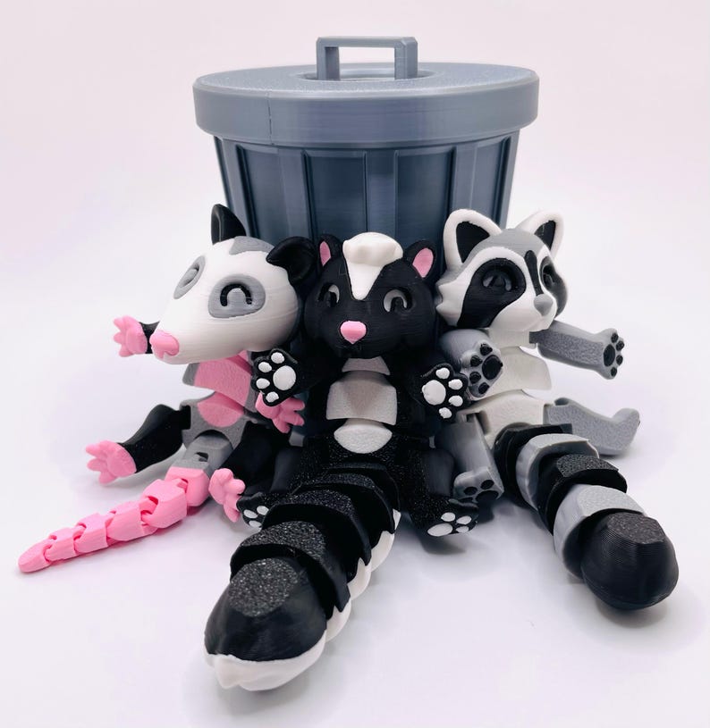 Op de afbeelding: Drie gearticuleerde dierenfiguren, een wasbeer, een stinkdier en een opossum, zijn geclusterd onder een grijze plastic prullenbak. De figuren zijn gemaakt van zwart, wit en roze plastic.