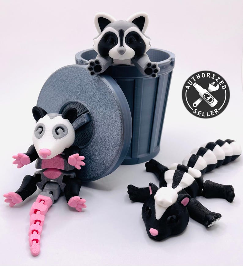 Op de afbeelding: Drie 3D-geprinte dierenfiguren: een wasbeer in een grijze prullenbak, een opossum met een roze staart en een stinkdier met een zwart-wit gestreepte staart.