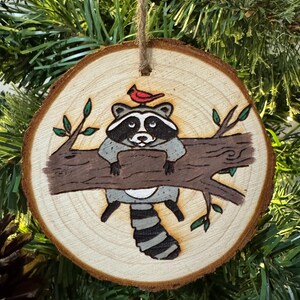 Op de afbeelding: Een houten kerstversiering met een handgeschilderde wasbeer met een rode kardinaal op zijn kop. De wasbeer zit op een tak met groene bladeren. De versiering is rond en heeft een rustieke houtnerf.