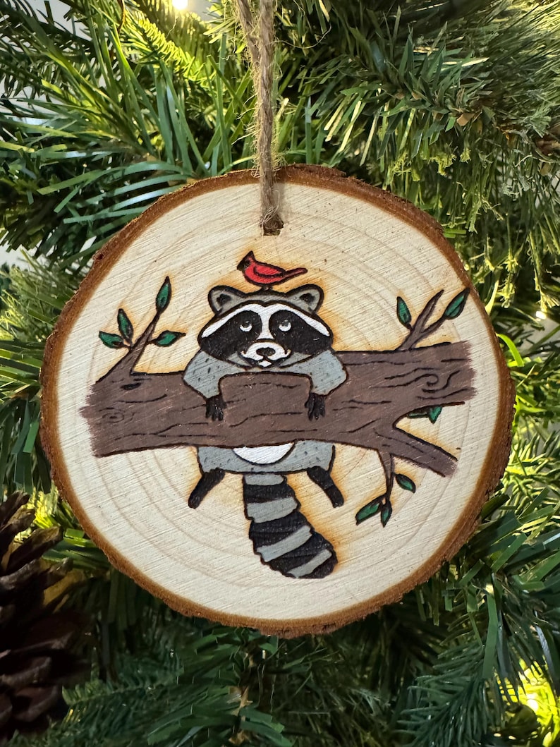 Op de afbeelding: Een houten kerstversiering met een handgeschilderde wasbeer met een rode kardinaal op zijn kop. De wasbeer zit op een tak met groene bladeren. De versiering is rond en heeft een rustieke houtnerf.