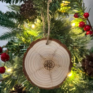 Op de afbeelding: Een houten kerstversiering met een natuurlijke houten afwerking en een cirkelvormig ontwerp. De versiering heeft een bruine touwhanger en een gemonogrammeerd "KP" in het midden.