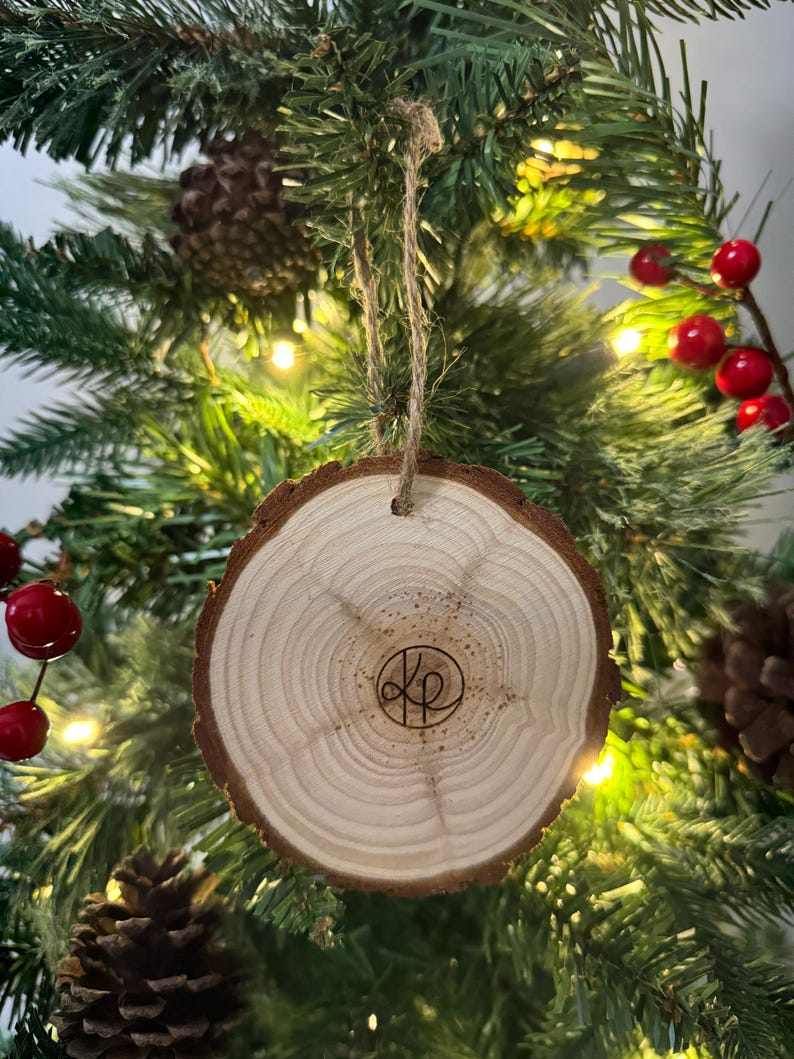 Op de afbeelding: Een houten kerstversiering met een natuurlijke houten afwerking en een cirkelvormig ontwerp. De versiering heeft een bruine touwhanger en een gemonogrammeerd "KP" in het midden.