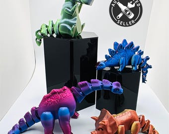 May include: Four colorful articulated dinosaur toys, each with a different color scheme and pose. The dinosaurs are made of plastic and have a segmented design. The dinosaurs are arranged on a white surface.
