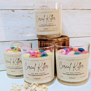 May include: Four glass jars filled with a white candle and colorful cereal-shaped decorations. The jars have labels that read "Mystic Healing Candle Co." and "Cereal Killer" with the website address "https://mystichealingus.etsy.com".