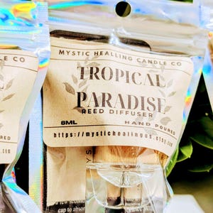 May include: An 8ml reed diffuser in a clear glass bottle, packaged in a holographic zip-lock pouch. The label reads Mystic Healing Candle Co, Tropical Paradise Reed Diffuser, Hand Poured, and includes the URL https://mystichealingus.etsy.com.