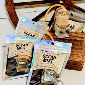 May include: A collection of clear glass car reed diffuser bottles with wooden caps and tan hanging cords, each packaged in a holographic resealable pouch labeled Mystic Healing Candle Co Ocean Mist Reed Diffuser Hand Poured 8ml.