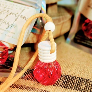 Puede incluir: Un ambientador para coche con líquido rojo dentro de una pequeña botella de vidrio redonda. Tiene una tapa de madera y está suspendido por un cordón beige. La etiqueta dice "Watermelon Splash".