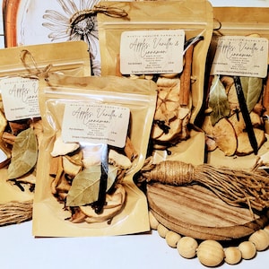 Puede incluir: Varias bolsas de plástico transparente llenas de rodajas de manzana secas, palitos de canela y otras especias. Cada bolsa tiene una etiqueta que dice "Apples, Vanilla & Cinnamon". Las bolsas están atadas con hilo y dispuestas con un elemento decorativo de madera.