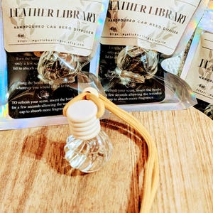 May include: Clear glass car reed diffuser with a wooden cap and a yellow cord. The packaging reads "Leather Library" and "Handpoured Car Reed Diffuser". The product is designed to refresh the scent in a car.