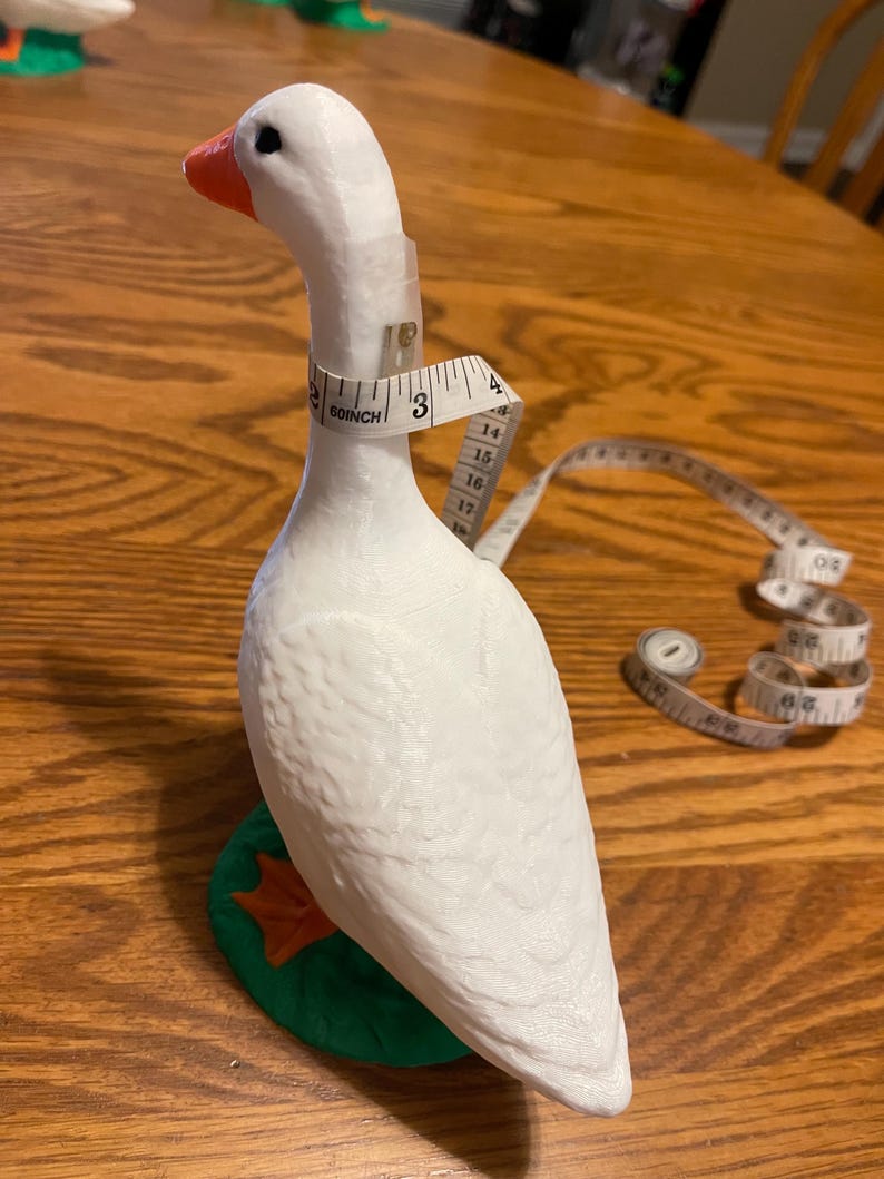 K&ouml;nnte beinhalten: Wei&szlig;e G&auml;nsefigur mit rotem Schnabel und gr&uuml;nem Sockel. Ein Ma&szlig;band ist um den Hals der Gans gelegt, mit einer Messung von 7,6 cm.