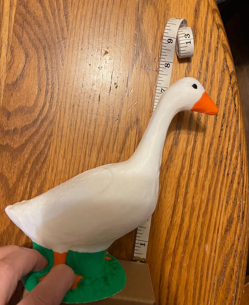 K&ouml;nnte beinhalten: Wei&szlig;e Keramik-G&auml;nsefigur mit orangefarbenem Schnabel und gr&uuml;nen F&uuml;&szlig;en. Die Gans steht auf einem gr&uuml;nen Sockel.