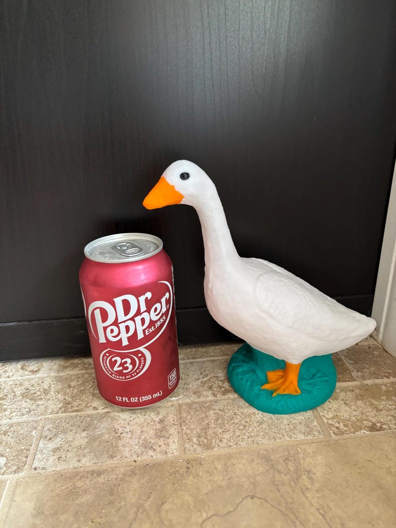 K&ouml;nnte beinhalten: Eine wei&szlig;e G&auml;nsefigur mit orangefarbenen F&uuml;&szlig;en und Schnabel, die auf einem t&uuml;rkisfarbenen Sockel steht. Die Gans steht neben einer roten Dose Dr Pepper mit dem Text "Dr Pepper Est. 1885" und "23" auf der Dose. Die Dose hat ein Fassungsverm&ouml;gen von 355 ml.