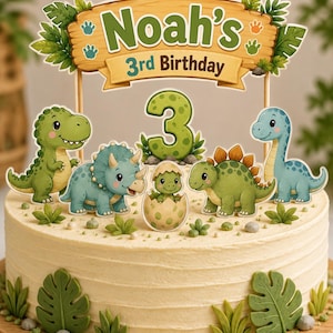 May include: A round cake with light-colored frosting, decorated with green fondant leaves and small rocks. A cake topper features a wooden-style sign reading Noah's 3rd Birthday, a large green number 3, and five cartoon dinosaur figures.