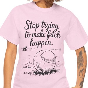 Może przedstawiać: Jasnoróżowa koszulka z czarnym napisem "Stop trying to make fetch happen". Grafika zawiera sylwetkę psa, dużą piłkę i trawę. Codzienna, swobodna odzież.