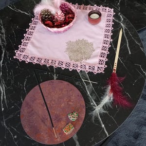 May include: A decorative arrangement on a black marble table. A pink basket holds pinecones and feathers. A pink doily with a floral design and a small candle holder are also present. An incense stick rests on a round, reddish-brown surface.