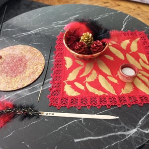 May include: A decorative arrangement on a black marble table. It includes a red and gold incense holder, a red placemat with gold leaf designs, a small bowl with pinecones, and a feather duster. A small candle sits on the placemat.