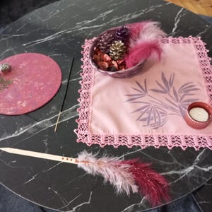 May include: A decorative arrangement on a black marble table. It includes a pink placemat with a lace border, a small candle holder, a bowl with feathers and pinecones, a round pink tray, incense sticks, and a feather wand.