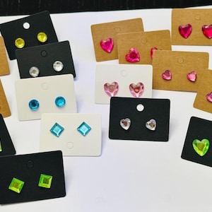 May include: A collection of stud earrings on display. The earrings feature various shapes, including hearts, squares, and circles, in colors like red, pink, green, blue, and clear. They are mounted on black, white, and brown earring display cards.