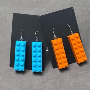 May include: Two pairs of earrings, each featuring a rectangular brick design. One pair is bright blue, the other is orange. The earrings have silver-colored hooks and are displayed on black cards against a gray background.