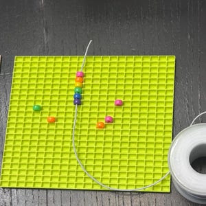 May include: A square lime green grid mat, a wooden ruler measuring up to 5 inches, a spool of white elastic cord, and several small multi-colored plastic beads. A string of beads is threaded onto a piece of the white elastic cord.