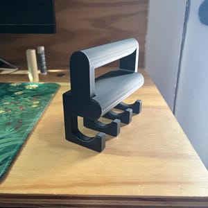 Puede incluir: Un colgador de pared de plástico negro con asa. El colgador tiene cuatro ganchos y está diseñado para sujetar objetos como llaves, toallas u otros objetos pequeños.