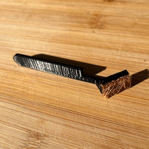 May include: A black cleaning tool with a small brown brush on the end. The tool is made of plastic and has a textured surface.