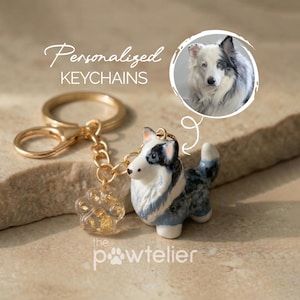 Op de afbeelding: Een gepersonaliseerde sleutelhanger met een keramieken hondenfiguur met blauwe, witte en zwarte markeringen. De sleutelhanger heeft een goudkleurige ring en een pootvormige bedel met goudvlokken. De tekst "Personalized KEYCHAINS" en "the Pawtelier" zijn zichtbaar.