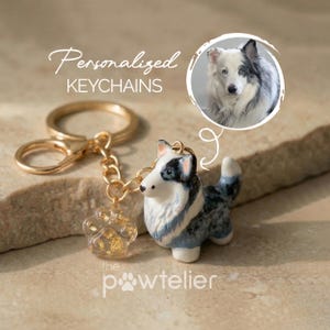 Puede incluir: Un llavero de metal dorado con una pequeña figura de perro de cerámica pintada a mano con marcas blue merle y un dije de resina transparente en forma de huella con copos de oro. El texto dice Personalized KEYCHAINS y the powtelier.