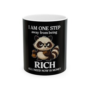 May include: A black and white ceramic mug with a white interior. The mug features a cartoon raccoon holding a money bag and a cookie, with the text "I AM ONE STEP away from being RICH ALL I NEED NOW IS MONEY."