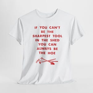 Può includere: T-shirt bianca con testo rosso: "SE NON PUOI ESSERE LO STRUMENTO PIÙ AFFILATO NEL CAPANNO, PUOI SEMPRE ESSERE LA ZAPPA". Il design include illustrazioni rosse di attrezzi da giardinaggio.