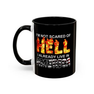 May include: Black and white ceramic mug with a black handle. The mug features the text "I'M NOT SCARED OF HELL I ALREADY LIVE IN AMERICA" in a bold, eye-catching design. The word "HELL" is in a fiery font.