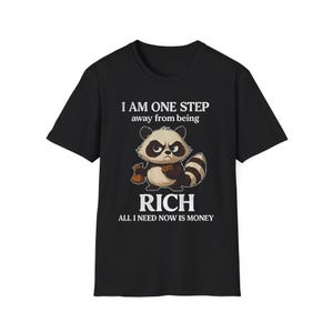 May include: Black t-shirt featuring a cartoon raccoon with a grumpy expression, holding a money bag. The text reads "I AM ONE STEP away from being RICH ALL I NEED NOW IS MONEY."