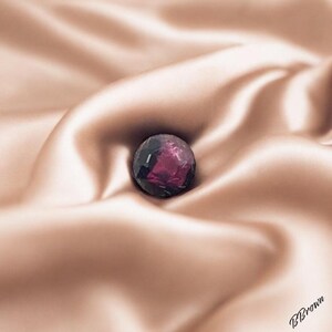 Peut inclure: Une pierre précieuse de couleur pourpre foncé avec des facettes, reposant sur un tissu satiné doux de couleur pêche.