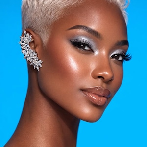 May include: A woman with short, platinum blonde hair and silver eyeshadow. She wears a sparkling ear cuff with clear crystals. Her skin is glowing against a bright blue background. The image is a beauty portrait.