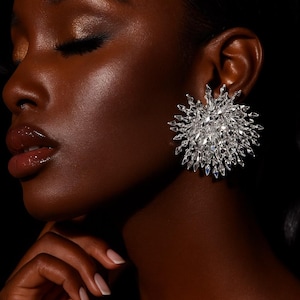 May include: A close-up of a person wearing a large, sparkling, silver-colored earring. The earring has a starburst design with many pointed, crystal-like elements. The person has dark skin and is wearing makeup. The background is black.