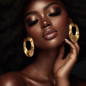 May include: A close-up of a person with dark skin, wearing gold earrings. The earrings are large, oval-shaped, and have a layered design. The person has closed eyes, with dark eye makeup and full lips. The background is dark.