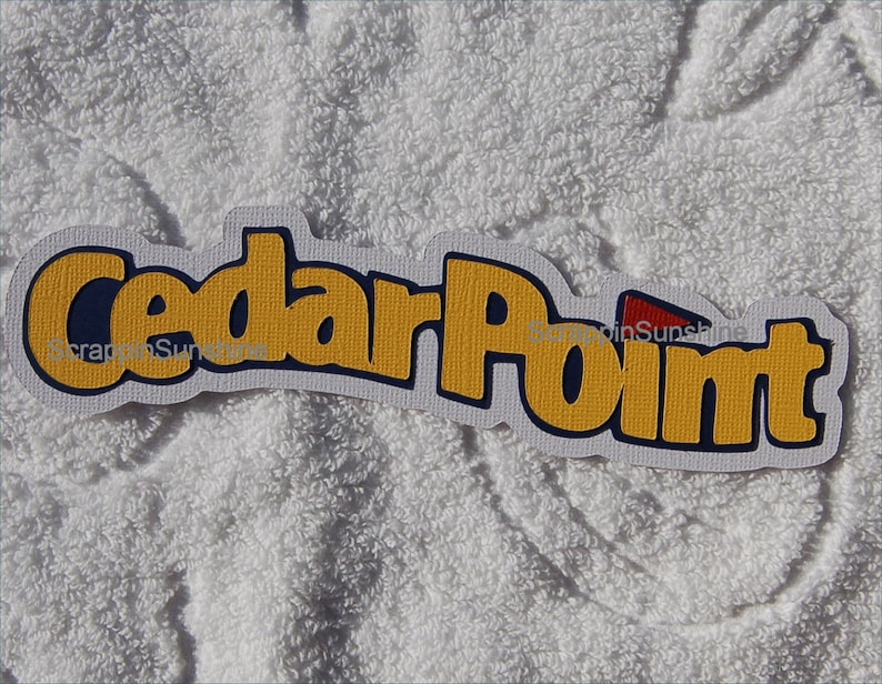 Op de afbeelding: Een wit, blauw en geel logo met de tekst "Cedar Point" in een vet, gestileerd lettertype. Het logo heeft een rode driehoek in de rechterbovenhoek.