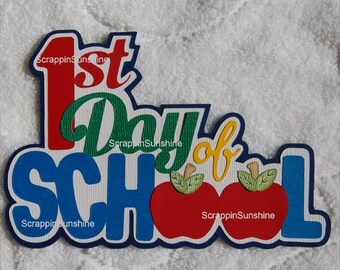 May include: A colorful paper craft with the words "1st Day of School" in red, green, yellow, and blue. The letters are outlined in navy blue and have a white background. Two red apples with green leaves are included in the design.