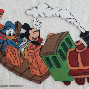 May include: A felt applique of Mickey Mouse, Donald Duck, and Goofy riding a train. The train is green and red with a brown wooden platform. The characters are smiling and waving. The train has a black smoke stack and a white cloud.