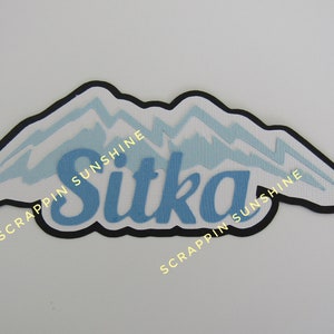 May include: A white and blue mountain range design with the word "Sitka" in blue lettering. The design is surrounded by a black border and the text "Scrappin Sunshine" is written on either side of the design.