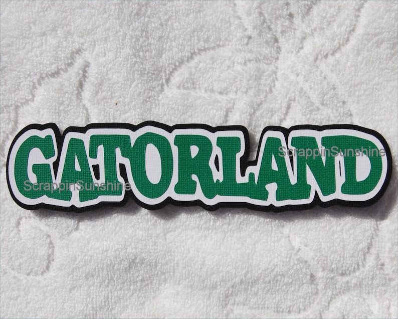 May include: A green and black paper craft cutout of the word "Gatorland" with a white background.