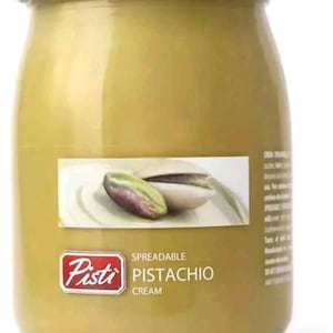 May include: A glass jar of pistachio cream spread with a white lid. The jar has a red label with the text "Pisti" and "Spreadable Pistachio Cream".