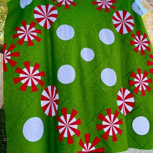 May include: A green quilt with a pattern of large white polka dots and red and white pinwheel designs. The quilt is made of a soft fabric and has a quilted texture. The overall theme is festive and whimsical.
