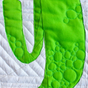 May include: Close-up of a lime green fabric letter "Y" with a bubble pattern, stitched onto a white quilted background. The letter is made of a solid green fabric with a decorative stitch design.