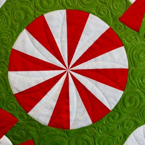 May include: Close-up of a quilted fabric with a repeating pattern of red and white peppermint candies on a green background. The candies have a spiral design. The green background has a swirl pattern.